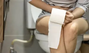 Femme assise sur des toilettes, tenant du papier toilette. Elle semble souffrir, peut-être d'une infection urinaire, et pense au yaourt comme remède.