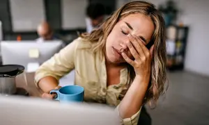 Femme au bureau, les yeux fermés, se frottant le visage, une tasse bleue à la main, signe d'une forte fatigue.