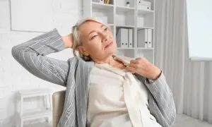 Femme d'âge mûr se rafraîchissant le cou, cherchant comment calmer les bouffées de chaleur. Elle est assise, la main derrière la tête, dans un bureau.