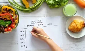 Planification d'un rééquilibrage alimentaire avec des légumes frais, un jus vert et un croissant. Une main écrit sur un plan de repas.