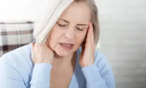 Femme aux cheveux blonds, les yeux fermés, se tenant la tête et le cou, visiblement souffrant de mal de tête tous les jours et fatigue.