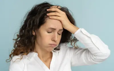 Femme aux cheveux bruns, le visage marqué par la fatigue mentale, une main sur le front, les yeux fermés.