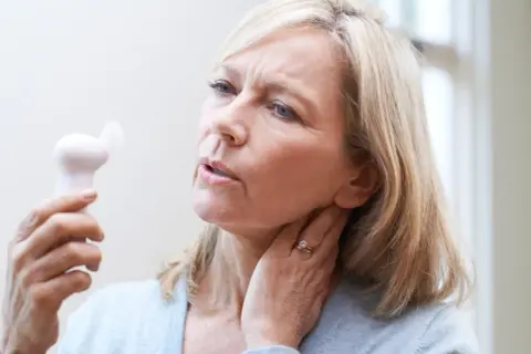 Femme blonde utilisant un petit ventilateur pour soulager les bouffées de chaleur de la préménopause. Elle a l'air inconfortable.