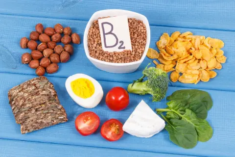 Assortiment d'aliments riches en vitamine B2 : sarrasin, noisettes, œuf, pain complet, tomates, brocolis, épinards, fromage et flocons de maïs.