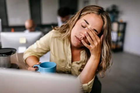 Femme au bureau, les yeux fermés, se frottant le visage, une tasse bleue à la main, signe d'une forte fatigue.