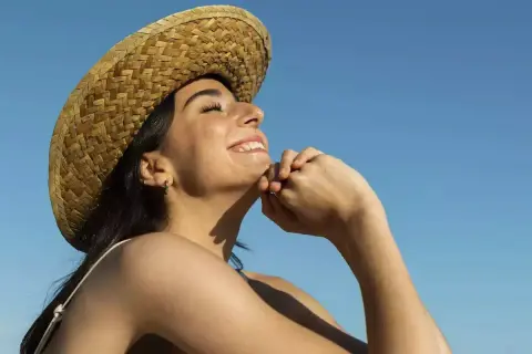 Jeune femme souriante sous un chapeau de paille, profitant du soleil pour sa vitamine D. Ciel bleu clair.
