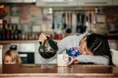 Une femme, accablée par une grosse fatigue soudaine, essaie de se servir un café. Un enfant la regarde, perplexe.