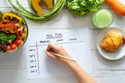 Planification d'un rééquilibrage alimentaire avec des légumes frais, un jus vert et un croissant. Une main écrit sur un plan de repas.
