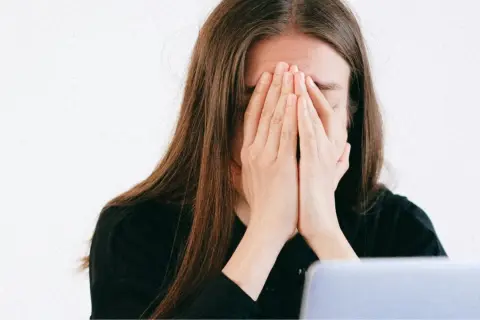 Une jeune femme, le visage caché par ses mains, exprime une **énorme fatigue** devant son ordinateur.