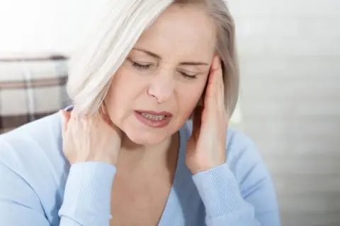 Femme aux cheveux blonds, les yeux fermés, se tenant la tête et le cou, visiblement souffrant de mal de tête tous les jours et fatigue.