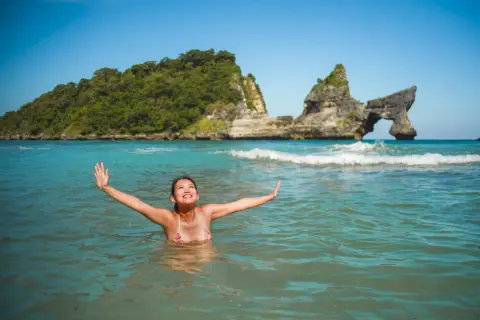 Une femme rayonnante profite des bienfaits de l'eau de mer dans un décor paradisiaque, bras ouverts vers le ciel bleu.