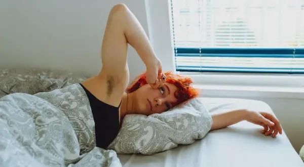 Jeune femme aux cheveux roux allongée dans son lit, le bras levé, l'air fatigué. Un manque de magnésium peut expliquer cette fatigue.