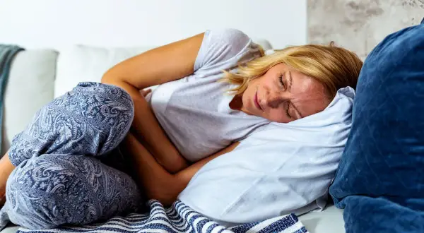 Femme blonde en pyjama, recroquevillée sur le canapé, se tenant le ventre. Elle semble souffrir de **difficulté à digérer**.