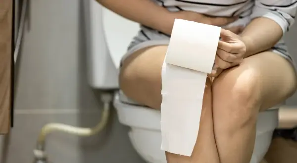 Femme assise sur des toilettes, tenant du papier toilette. Elle semble souffrir, peut-être d'une infection urinaire, et pense au yaourt comme remède.