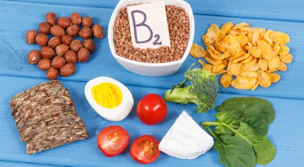 Assortiment d'aliments riches en vitamine B2 : sarrasin, noisettes, œuf, pain complet, tomates, brocolis, épinards, fromage et flocons de maïs.