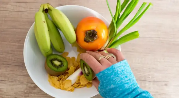 Bol de fruits avec banane, kaki, kiwi et céréales. Une main prépare une collation saine pour aider à prévenir une infection urinaire.