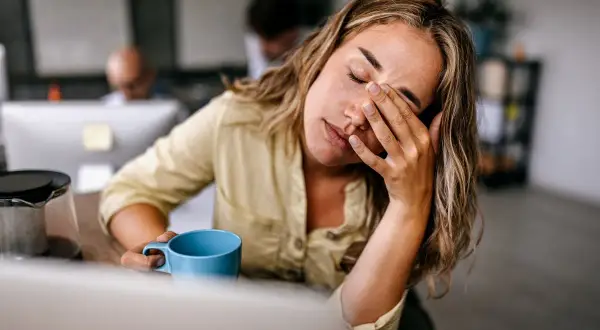 Femme au bureau, les yeux fermés, se frottant le visage, une tasse bleue à la main, signe d'une forte fatigue.
