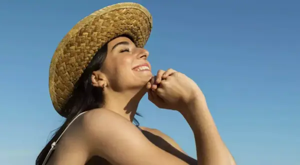 Jeune femme souriante sous un chapeau de paille, profitant du soleil pour sa vitamine D. Ciel bleu clair.