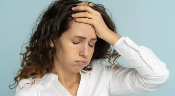 Femme aux cheveux bruns, le visage marqué par la fatigue mentale, une main sur le front, les yeux fermés.