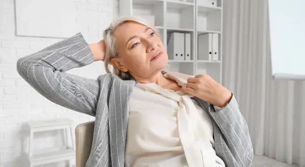 Femme d'âge mûr se rafraîchissant le cou, cherchant comment calmer les bouffées de chaleur. Elle est assise, la main derrière la tête, dans un bureau.