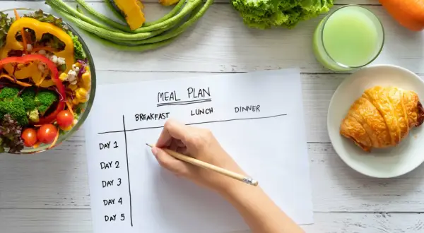 Planification d'un rééquilibrage alimentaire avec des légumes frais, un jus vert et un croissant. Une main écrit sur un plan de repas.