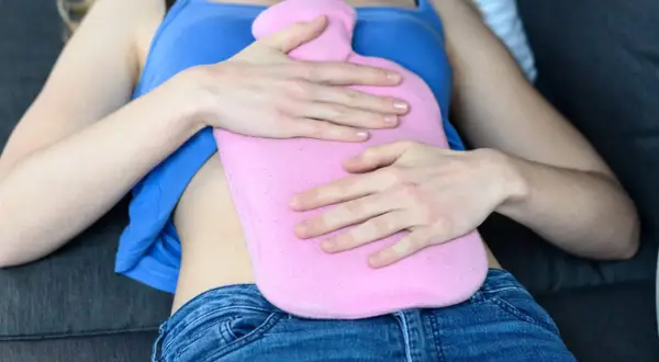 Une femme se tient une bouillotte rose sur le ventre, un remède de grand-mère pour une règle très douloureuse.