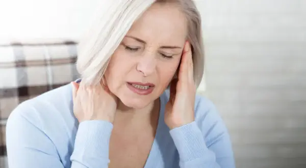 Femme aux cheveux blonds, les yeux fermés, se tenant la tête et le cou, visiblement souffrant de mal de tête tous les jours et fatigue.