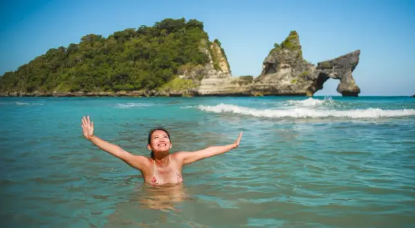 Une femme rayonnante profite des bienfaits de l'eau de mer dans un décor paradisiaque, bras ouverts vers le ciel bleu.