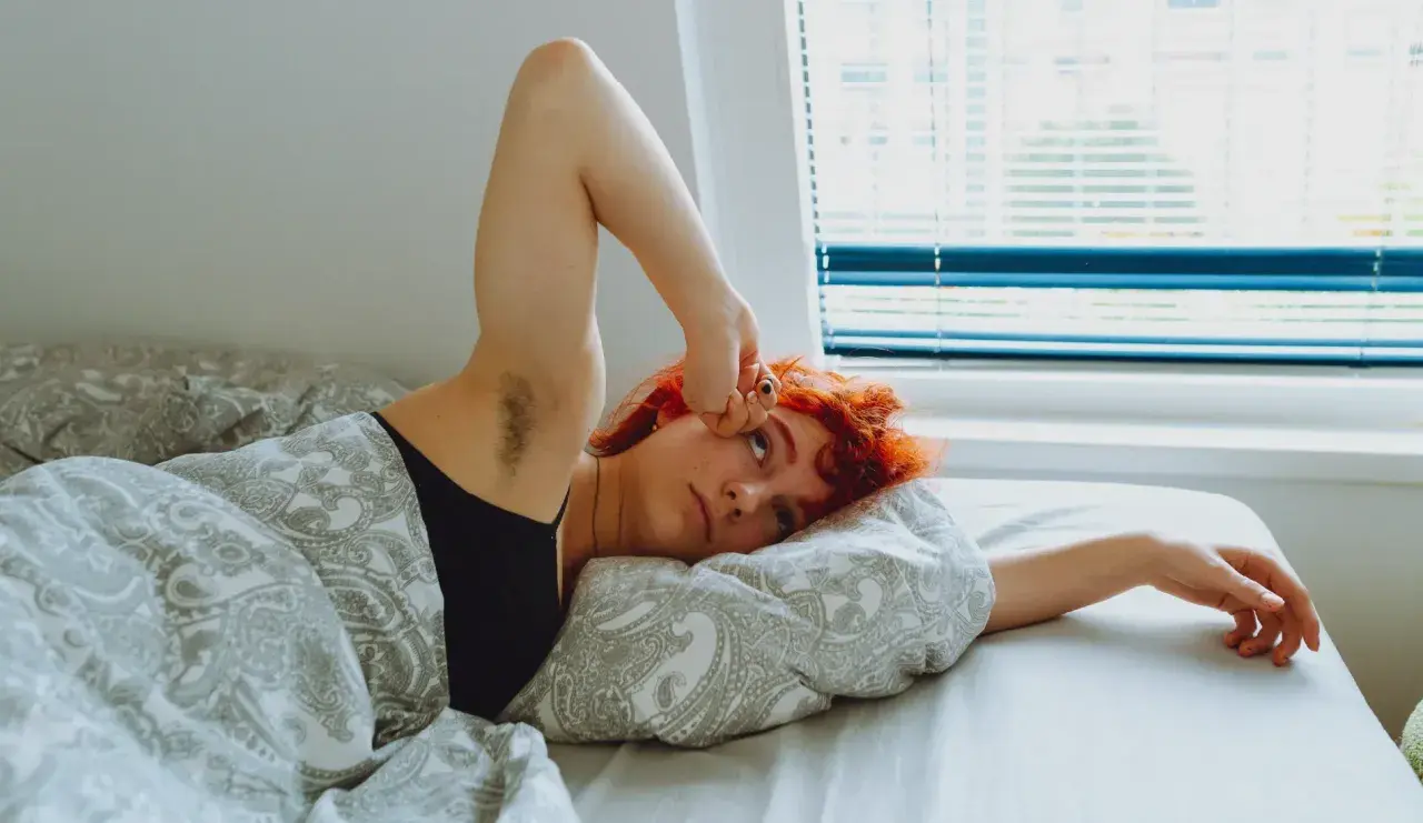 Jeune femme aux cheveux roux allongée dans son lit, le bras levé, l'air fatigué. Un manque de magnésium peut expliquer cette fatigue.