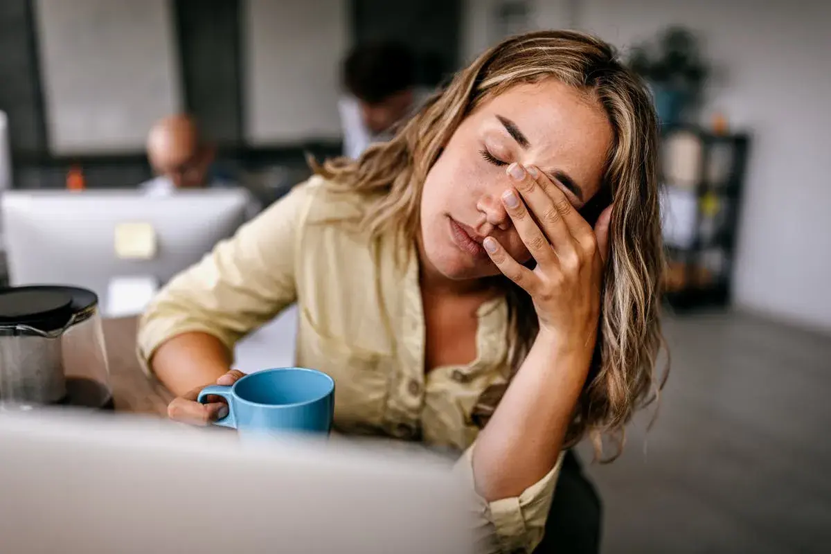 Femme au bureau, les yeux fermés, se frottant le visage, une tasse bleue à la main, signe d'une forte fatigue.