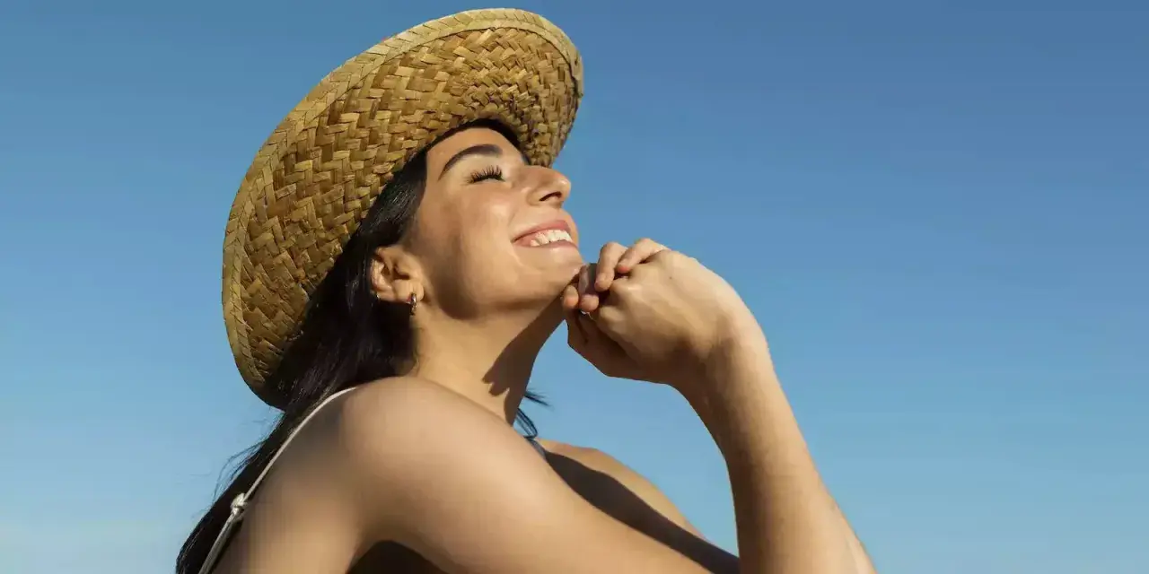 Jeune femme souriante sous un chapeau de paille, profitant du soleil pour sa vitamine D. Ciel bleu clair.