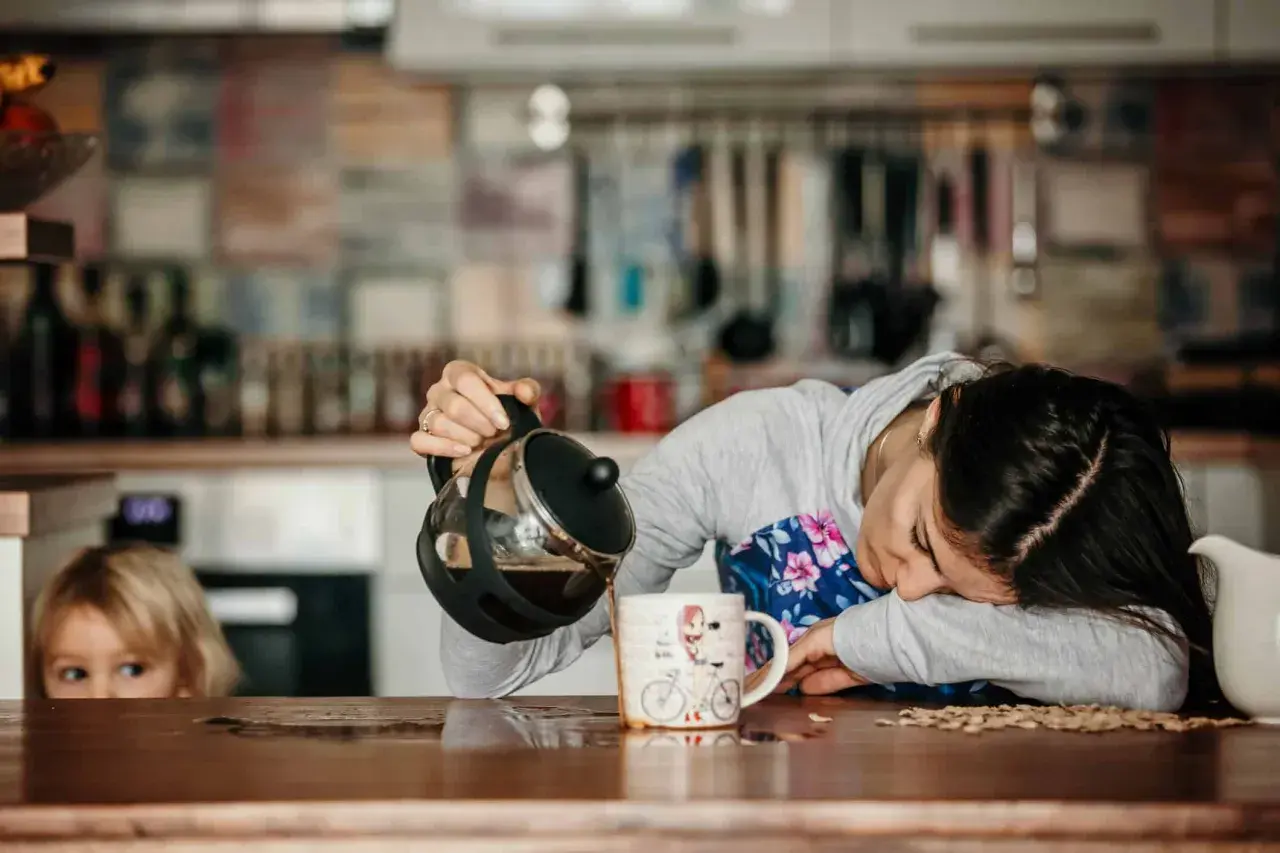 Une femme, accablée par une grosse fatigue soudaine, essaie de se servir un café. Un enfant la regarde, perplexe.