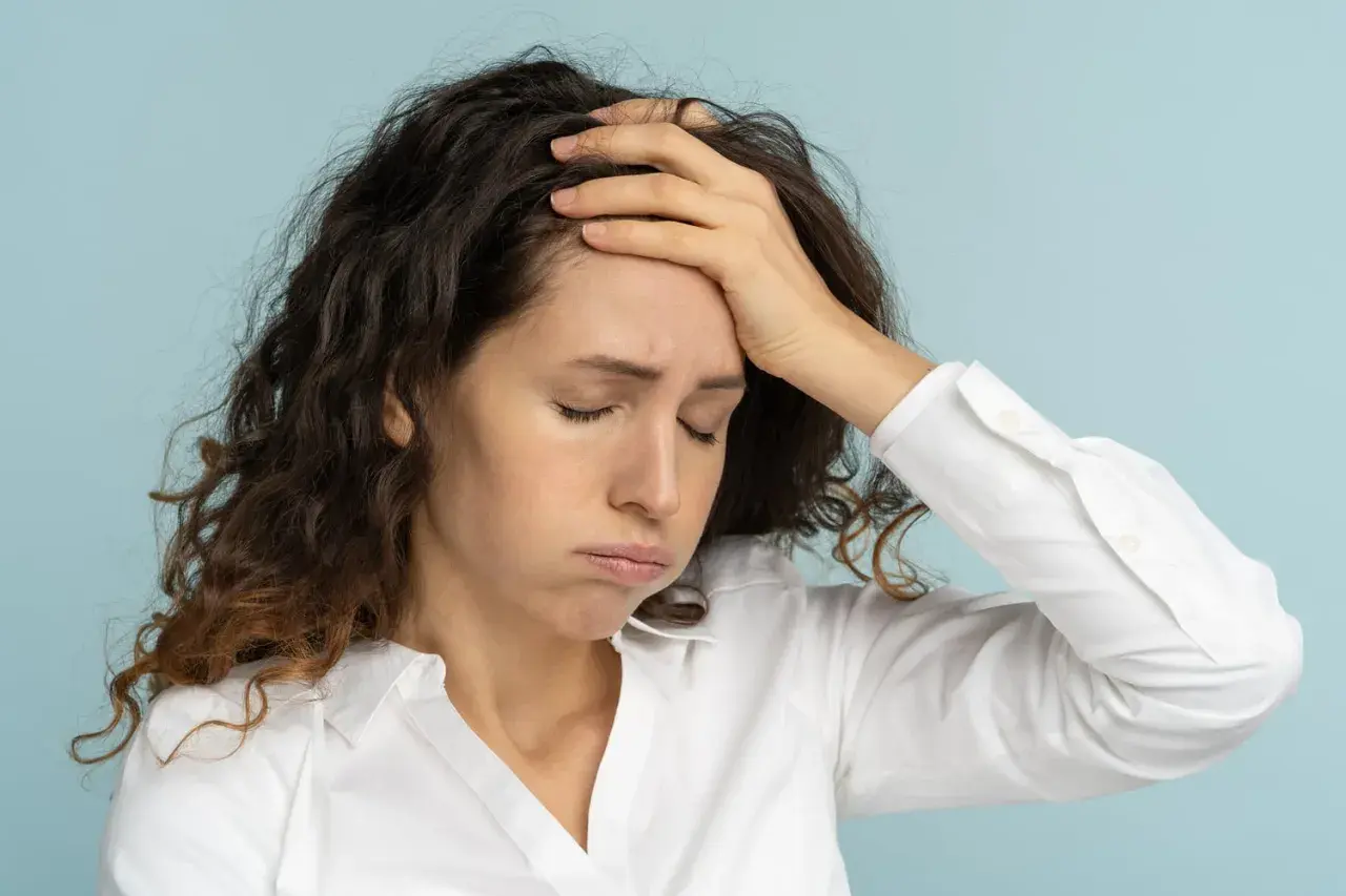 Femme aux cheveux bruns, le visage marqué par la fatigue mentale, une main sur le front, les yeux fermés.