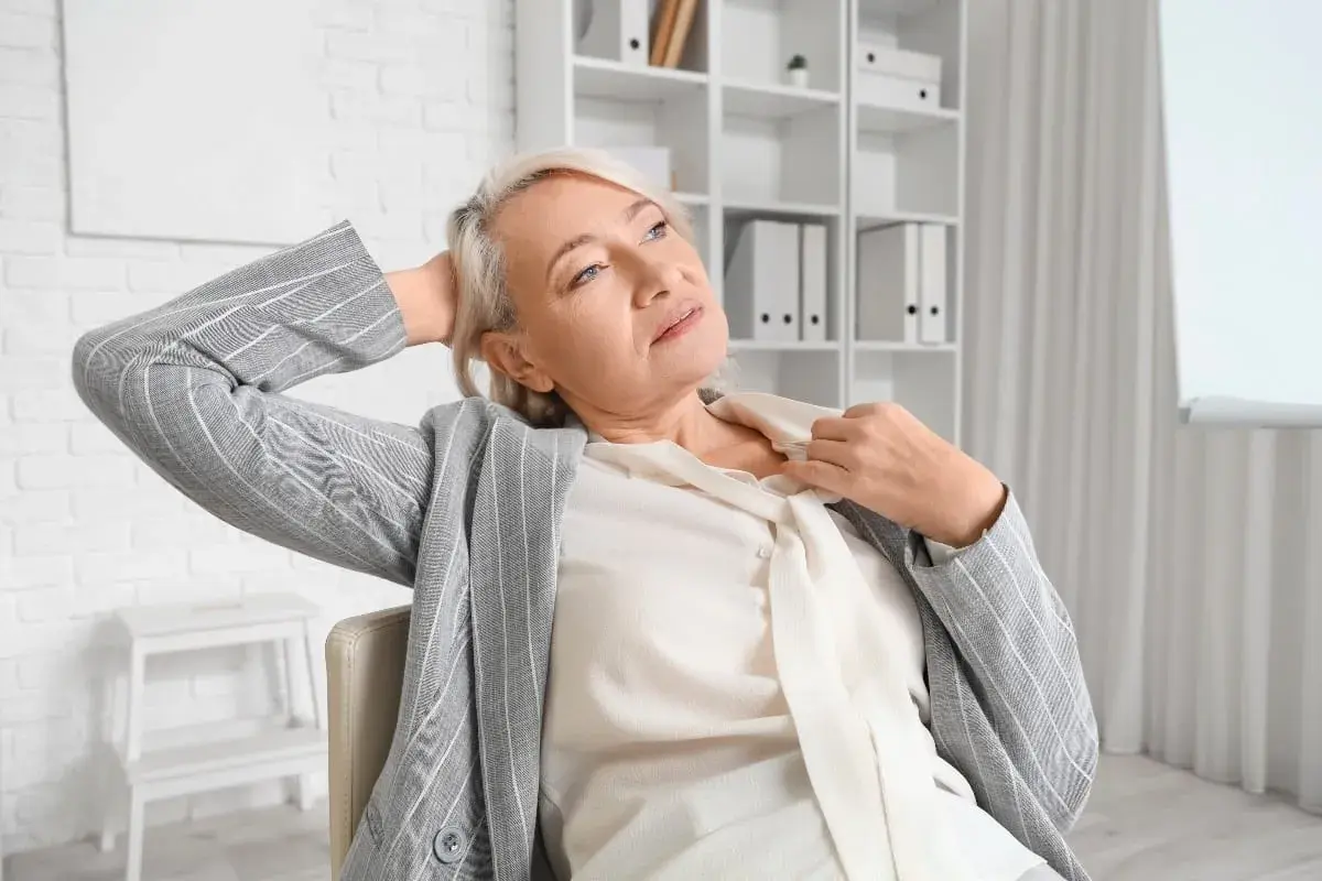Femme d'âge mûr se rafraîchissant le cou, cherchant comment calmer les bouffées de chaleur. Elle est assise, la main derrière la tête, dans un bureau.