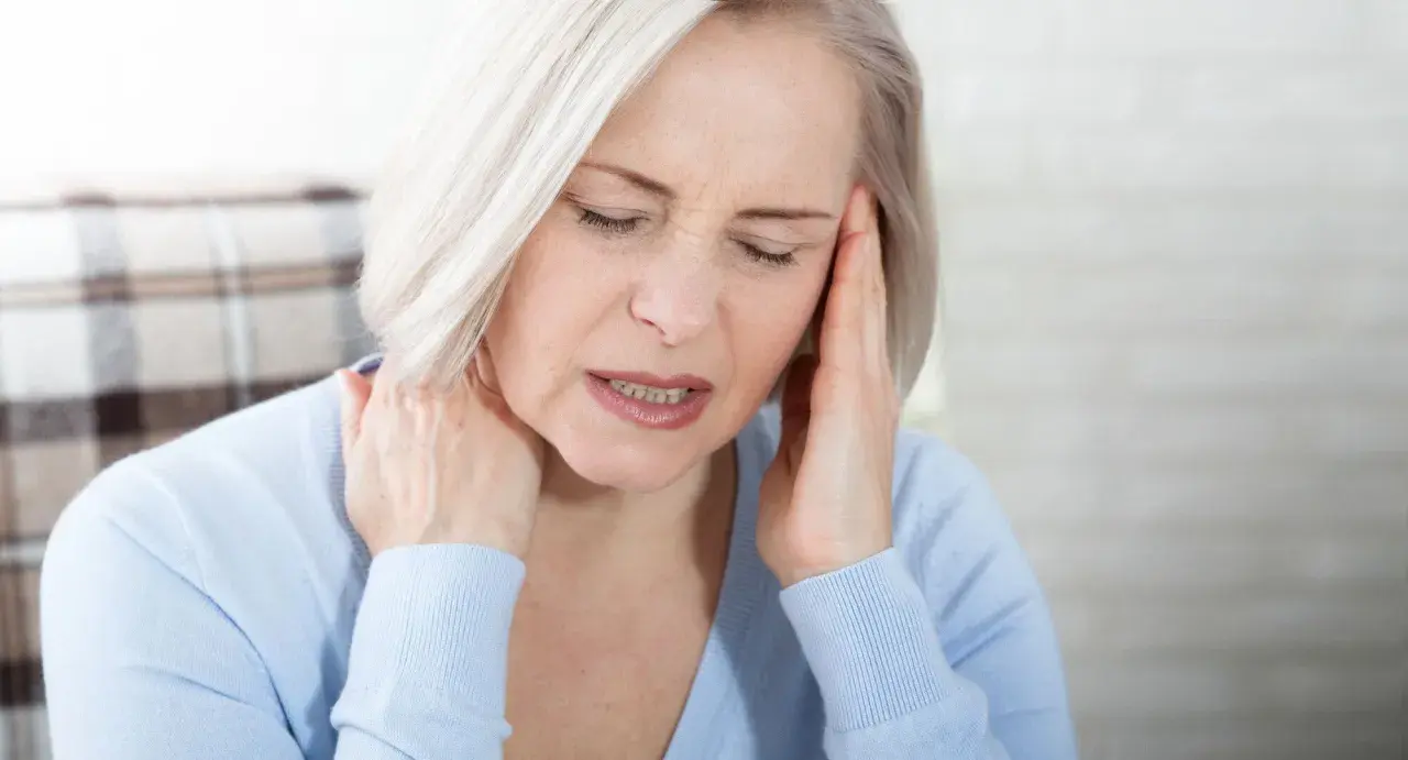 Femme aux cheveux blonds, les yeux fermés, se tenant la tête et le cou, visiblement souffrant de mal de tête tous les jours et fatigue.