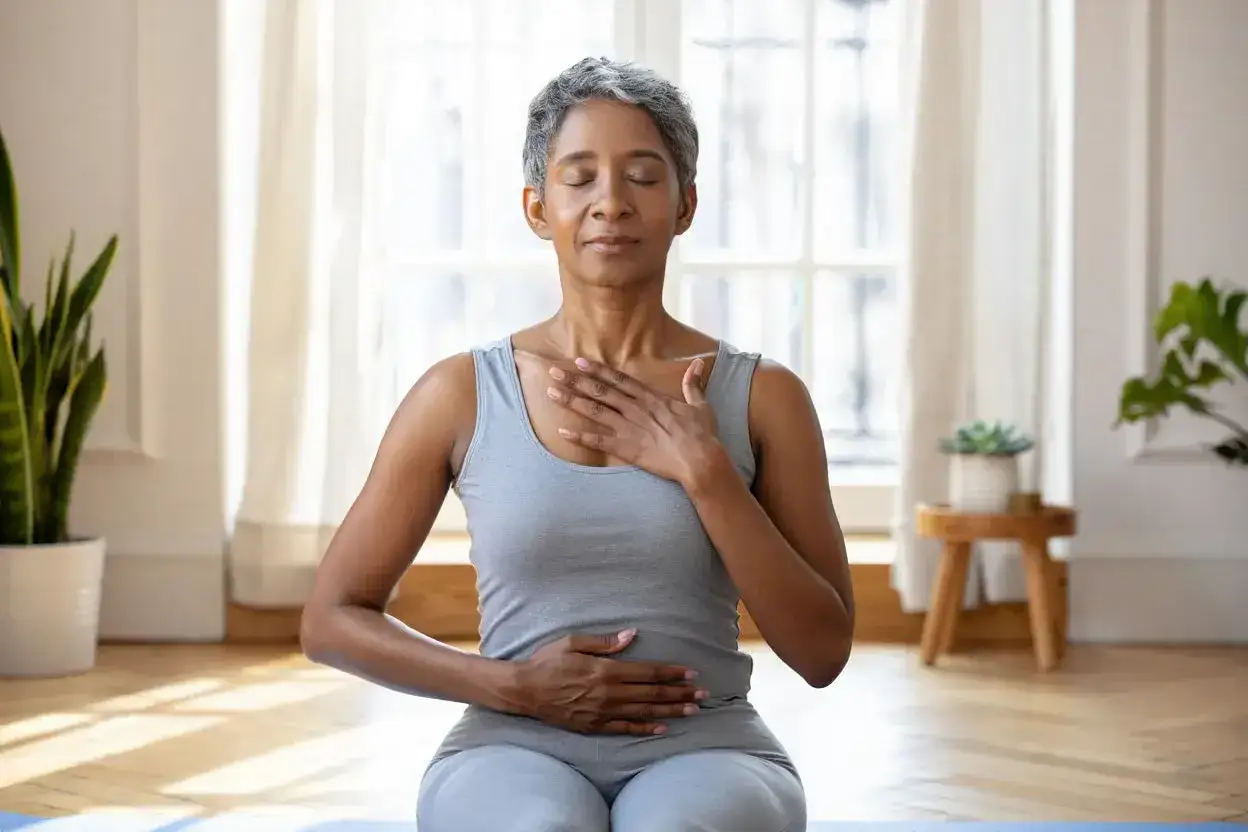 Femme pratiquant la respiration profonde pour faire baisser le cortisol. Mains sur la poitrine et le ventre, yeux fermés, dans une pièce lumineuse.