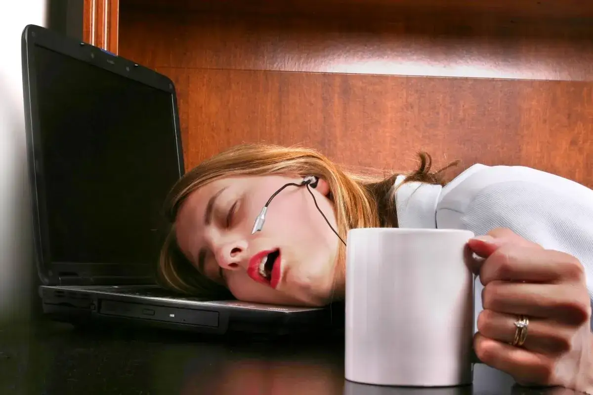Une jeune femme s'est endormie sur son ordinateur portable, une tasse de café à la main, signe d'une forte fatigue.