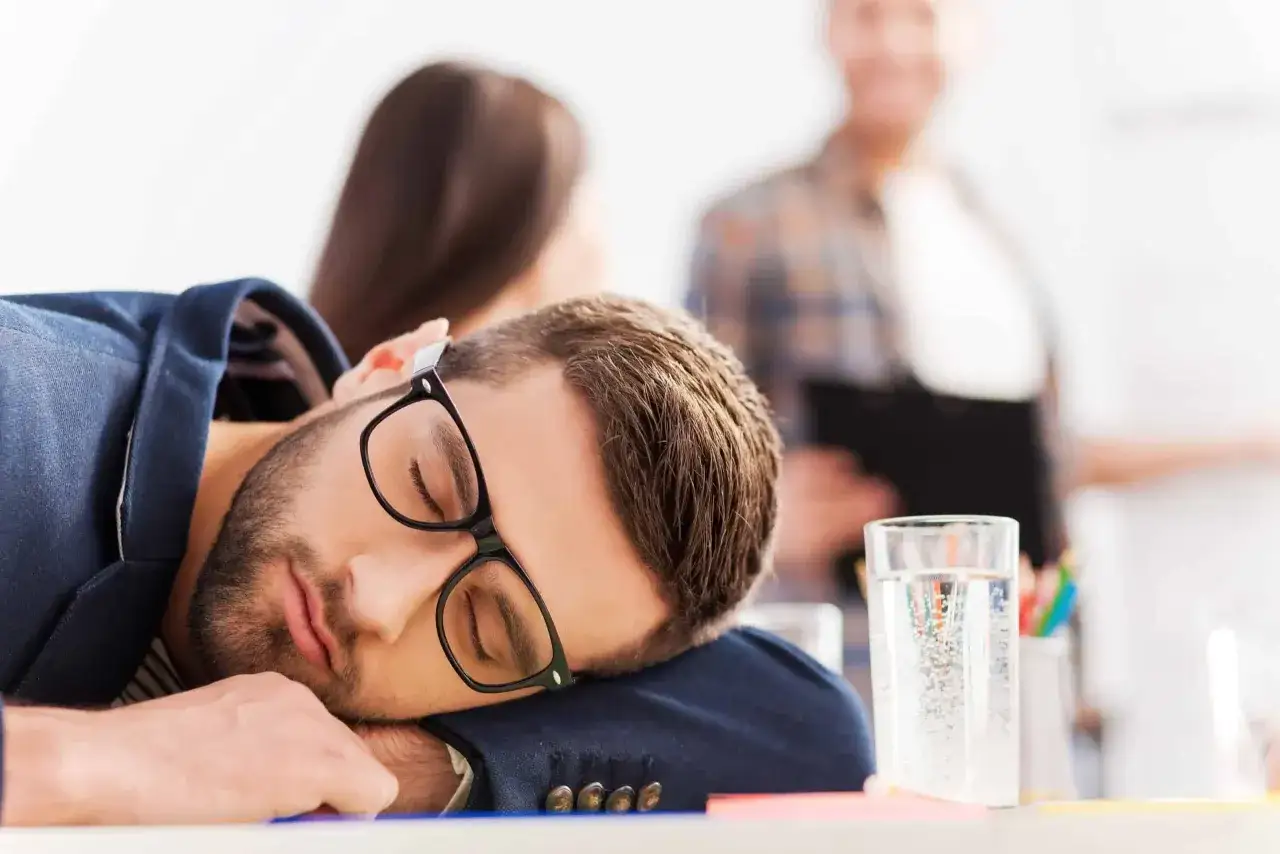 Un homme dort sur son bureau, lunettes sur le nez. Il montre bien que peut cacher une grande fatigue.