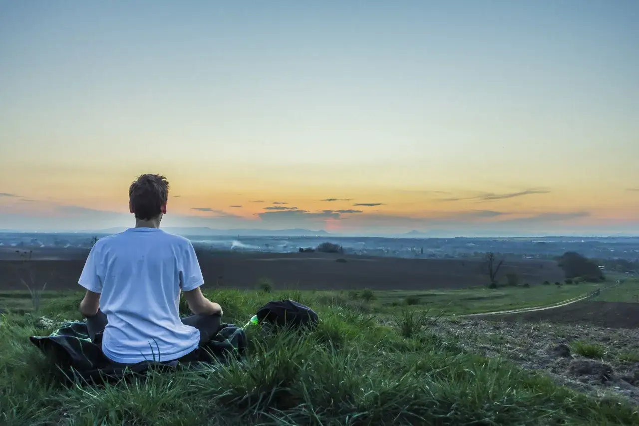Un jeune homme en t-shirt blanc pratique la m&eacute;ditation assis sur une couverture, contemplant un paysage rural au coucher du soleil.