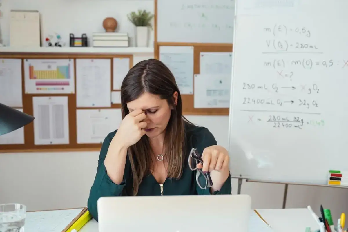 Une enseignante, visiblement épuisée, se frotte les tempes, un signe de stress symptomatique. Elle tient des lunettes à la main, devant un tableau rempli d'équations chimiques.