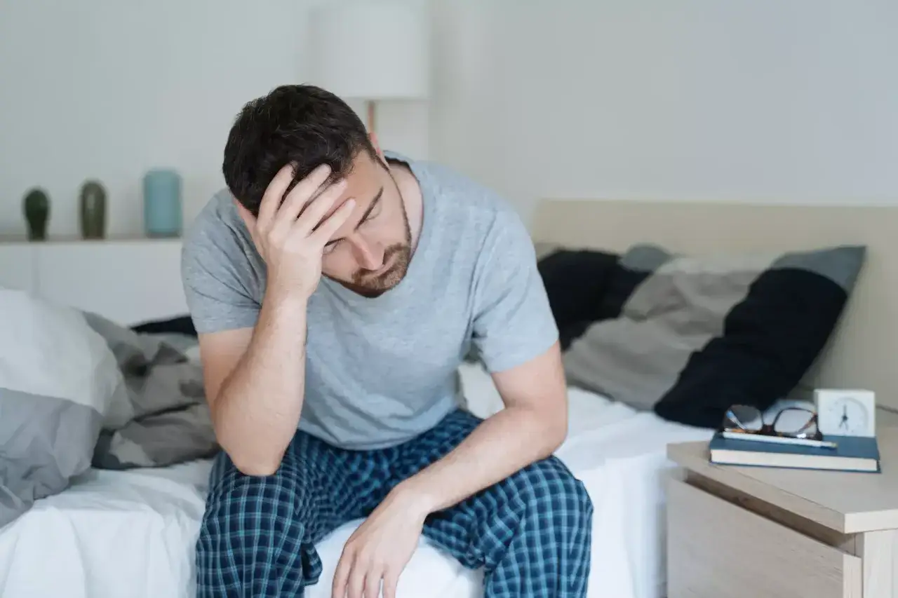 Homme assis sur son lit, la t&ecirc;te dans les mains, exprimant une profonde fatigue et un manque d'&eacute;nergie.