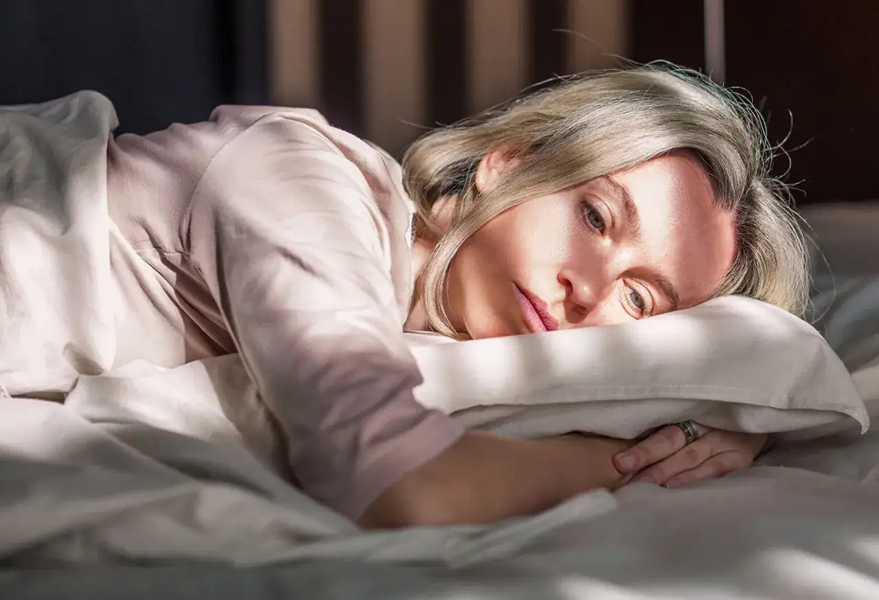 Femme au lit, le visage marqué par les bouffées de chaleur et la fatigue intense, sous un rayon de soleil.