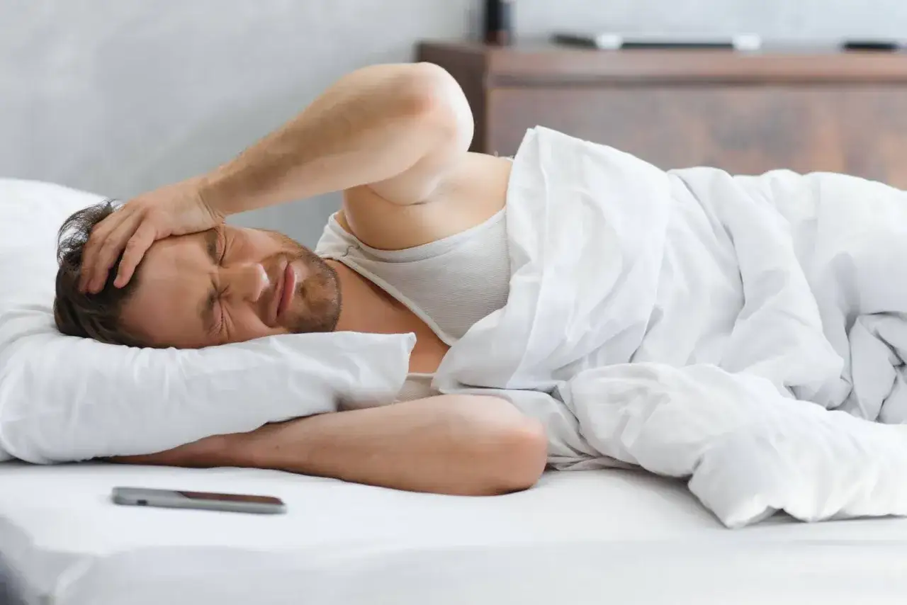 Un homme se réveille dans son lit, la main sur la tête, visiblement en proie à un épuisement total.