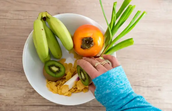 Bol de fruits avec banane, kaki, kiwi et céréales. Une main prépare une collation saine pour aider à prévenir une infection urinaire.