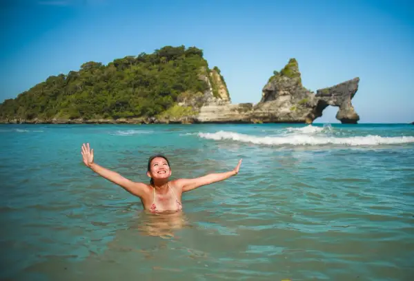 Une femme rayonnante profite des bienfaits de l'eau de mer dans un décor paradisiaque, bras ouverts vers le ciel bleu.